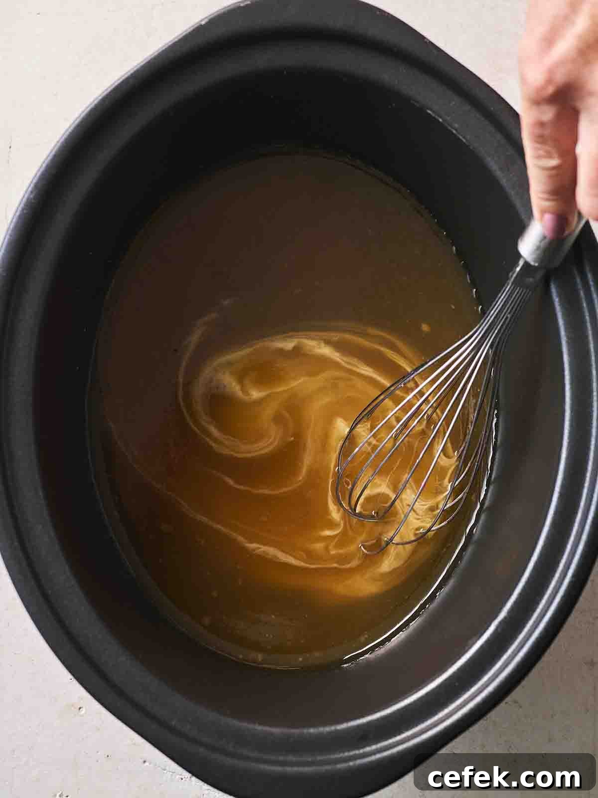 A slow cooker pan with gravy in the bottom, with thickening cornflour stirring in with a whisk, for the recipe Slow Cooker Lamb Leg.
