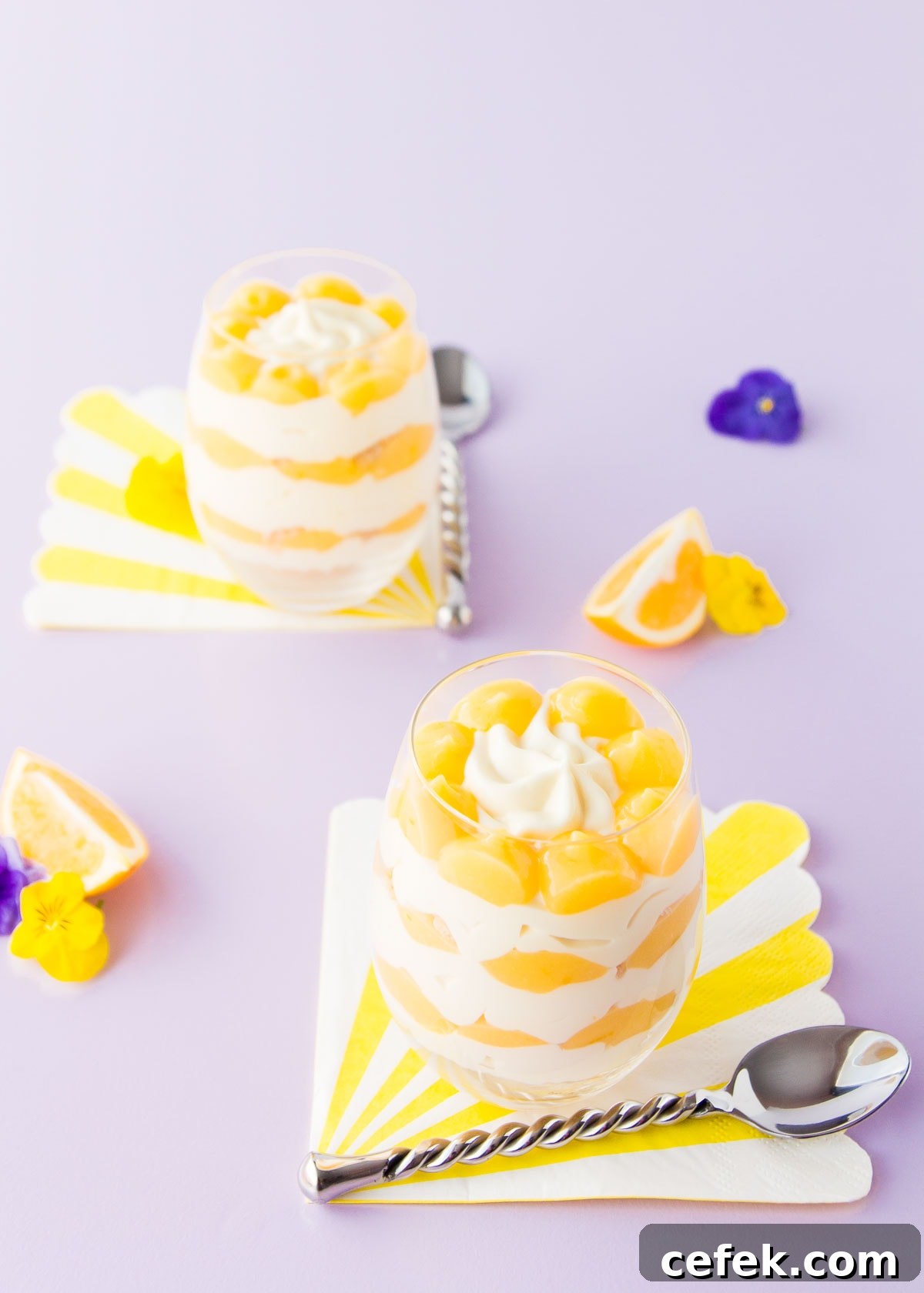 Two lemon cheesecake cups, on yellow and white striped napkins, on a pale purple background.