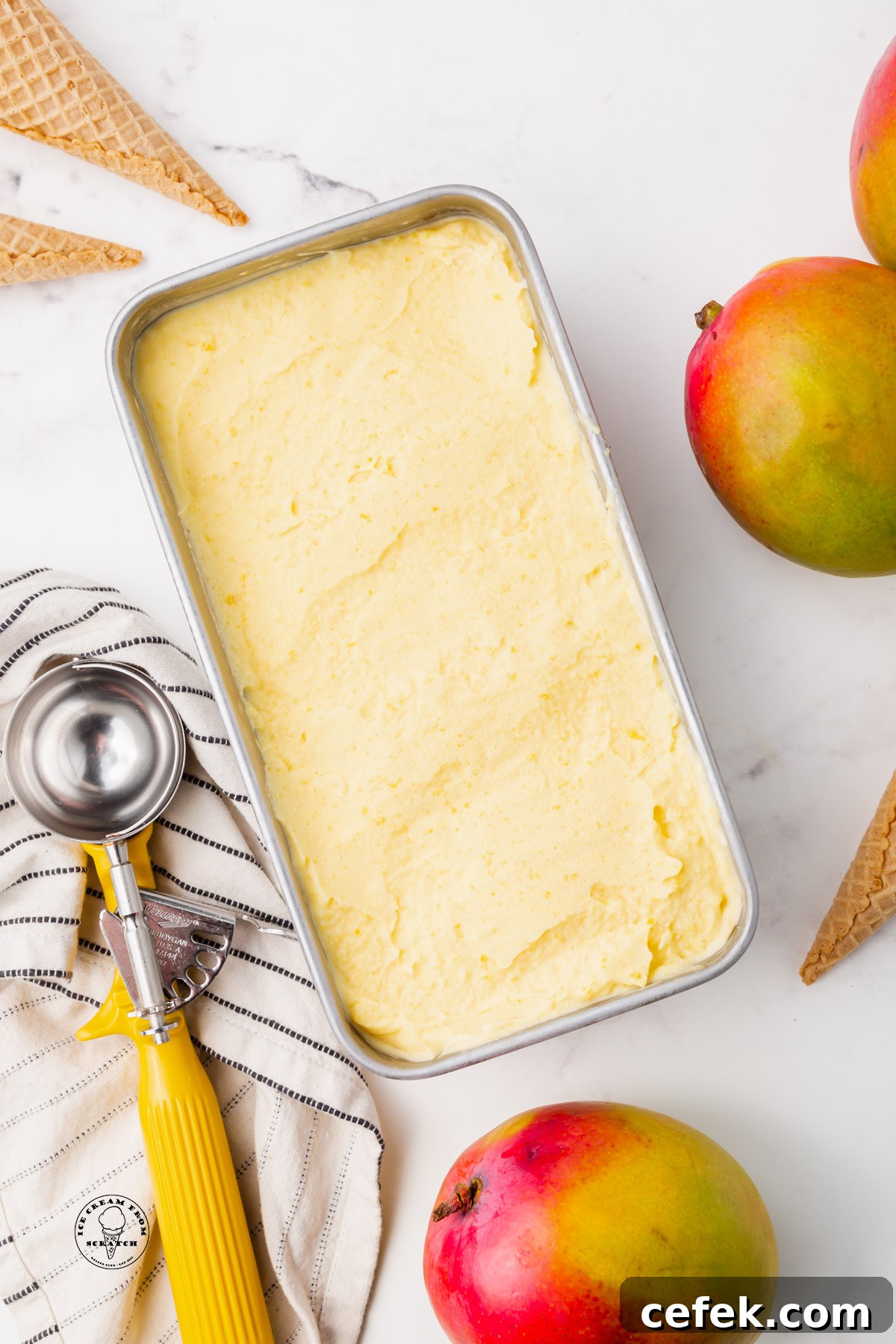 a loaf pan filled with creamy mango gelato. Next to the pan are fresh mangoes and a yellow metal ice cream scoop.