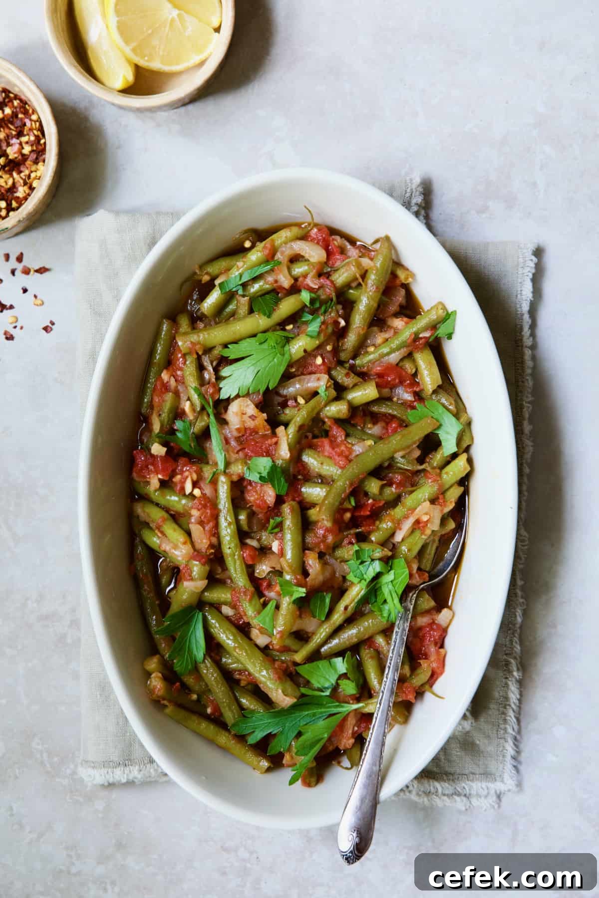 Mediterranean Braised Green Beans with Tomatoes in oval serving dish with serving spoon garnished with fresh parsley.