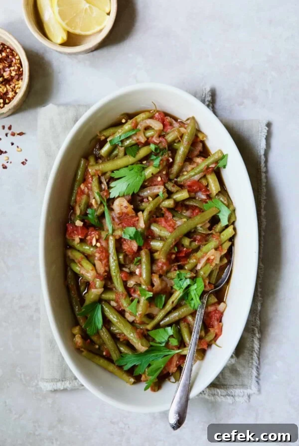 Mediterranean Braised Green Beans with Tomatoes in oval serving dish with serving spoon garnished with fresh parsley.