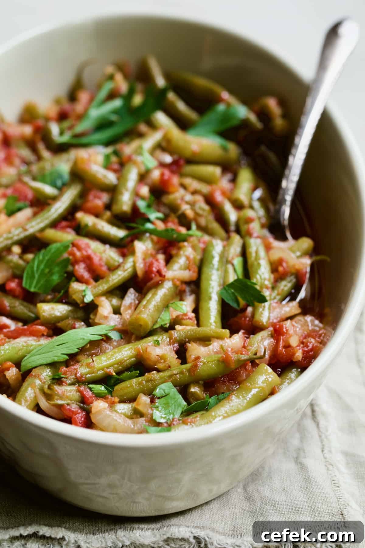 Mediterranean Braised Green Beans with Tomatoes in serving dish with serving spoon on beige fringed napkin.
