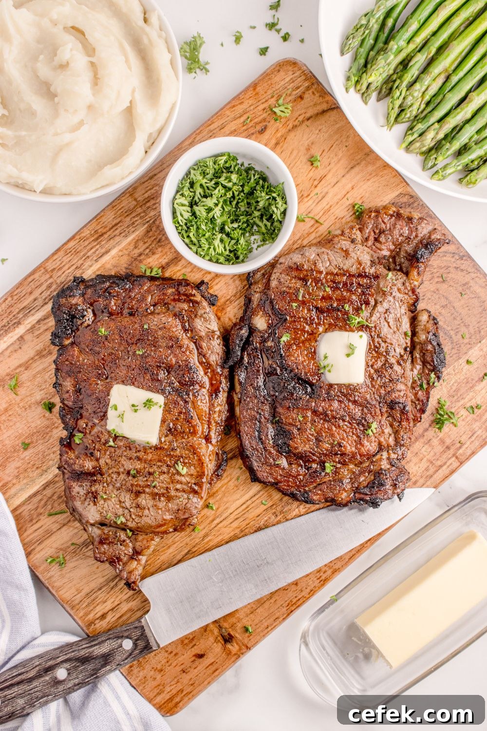 Grilled Ribeye Steaks on a cutting board with slabs of butter and bowl of parsley to the side 