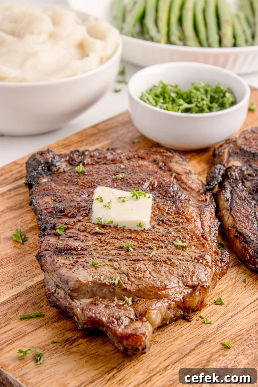 Grilled Ribeye Steak on cutting board with a slab of butter on top of steak and a bowl of parsley to side 
