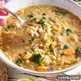 A white bowl of creamy chicken orzo soup with someone scooping up a spoonful, and with ingredients scattered around it.