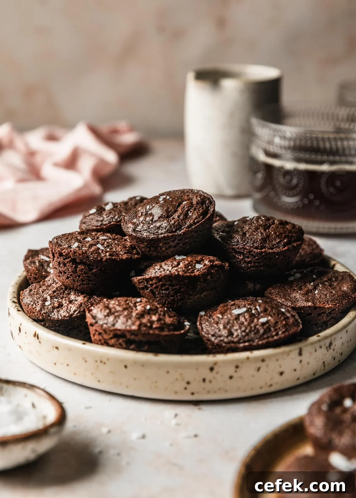 A tan plate of brownie bites on a tan counter next to a pink linen, white cream container, and cup of coffee.