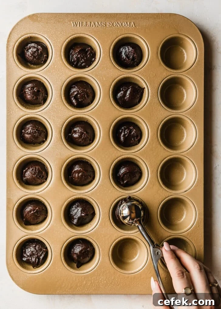 A hand using a scoop to divide brownie bites into a gold muffin pan on a tan counter.