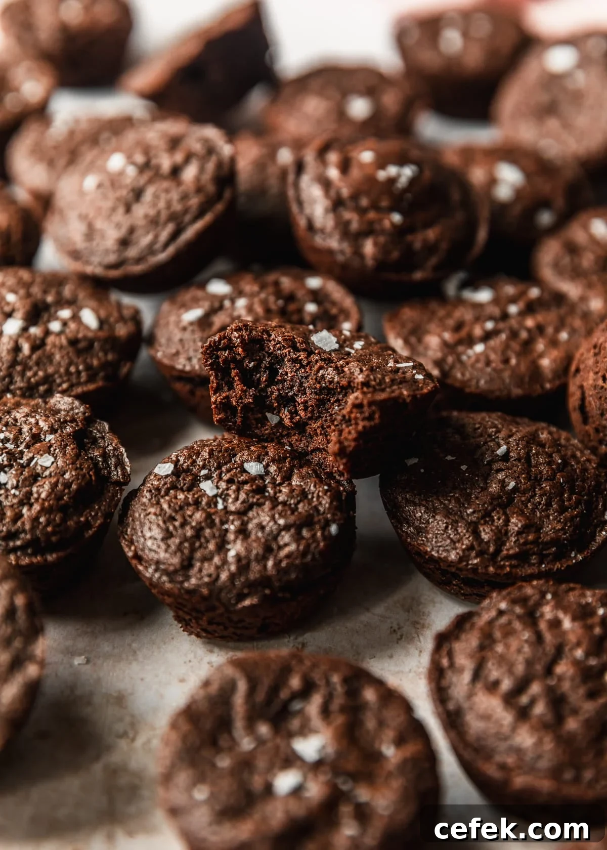 Rows and stacks of mini brownie bites with flaky salt on a tan counter.