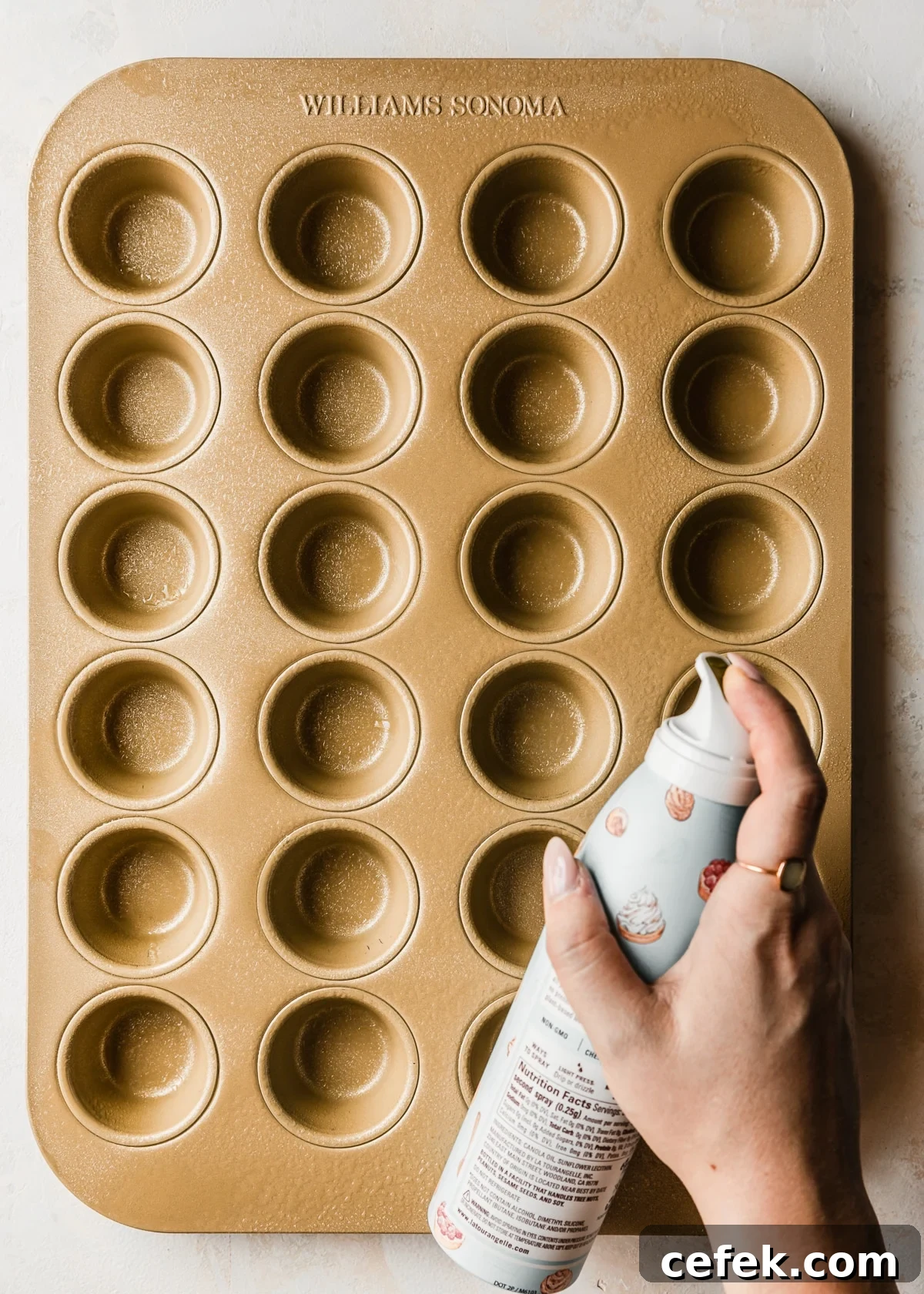 A hand spraying baking spray on a gold mini muffin tin on a tan counter.