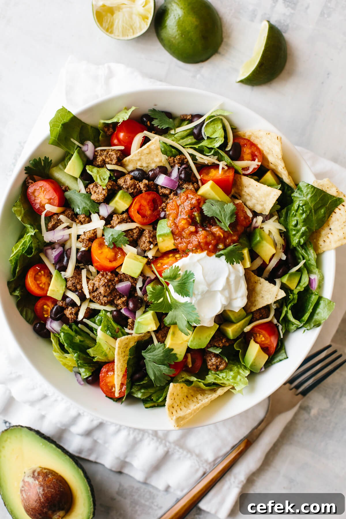 Taco salad in a white bowl on top of a napkin, with limes and half an avocado next to the bowl.
