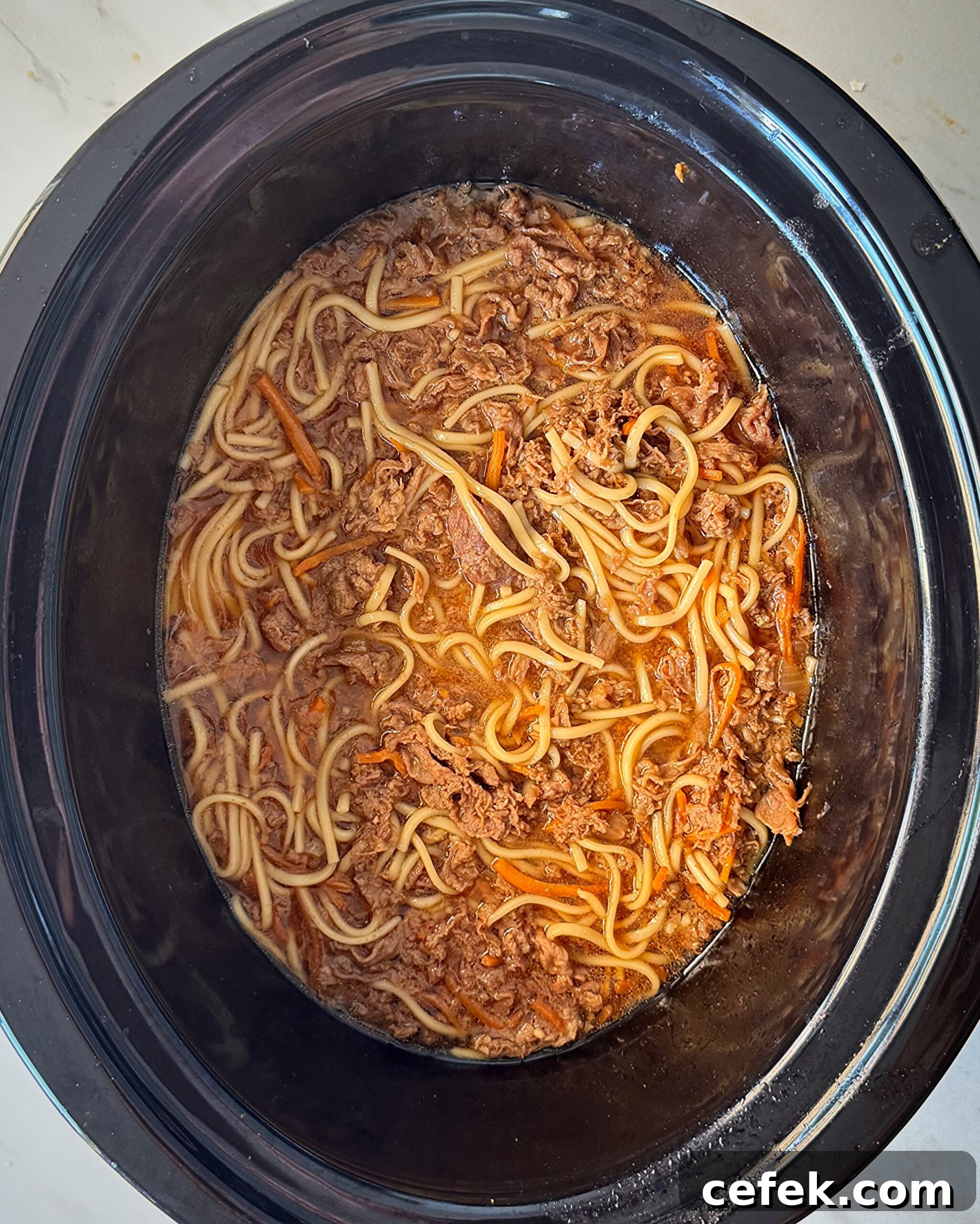 overhead shot of uncooked beef udon soup in a black slow cooker with noodles added