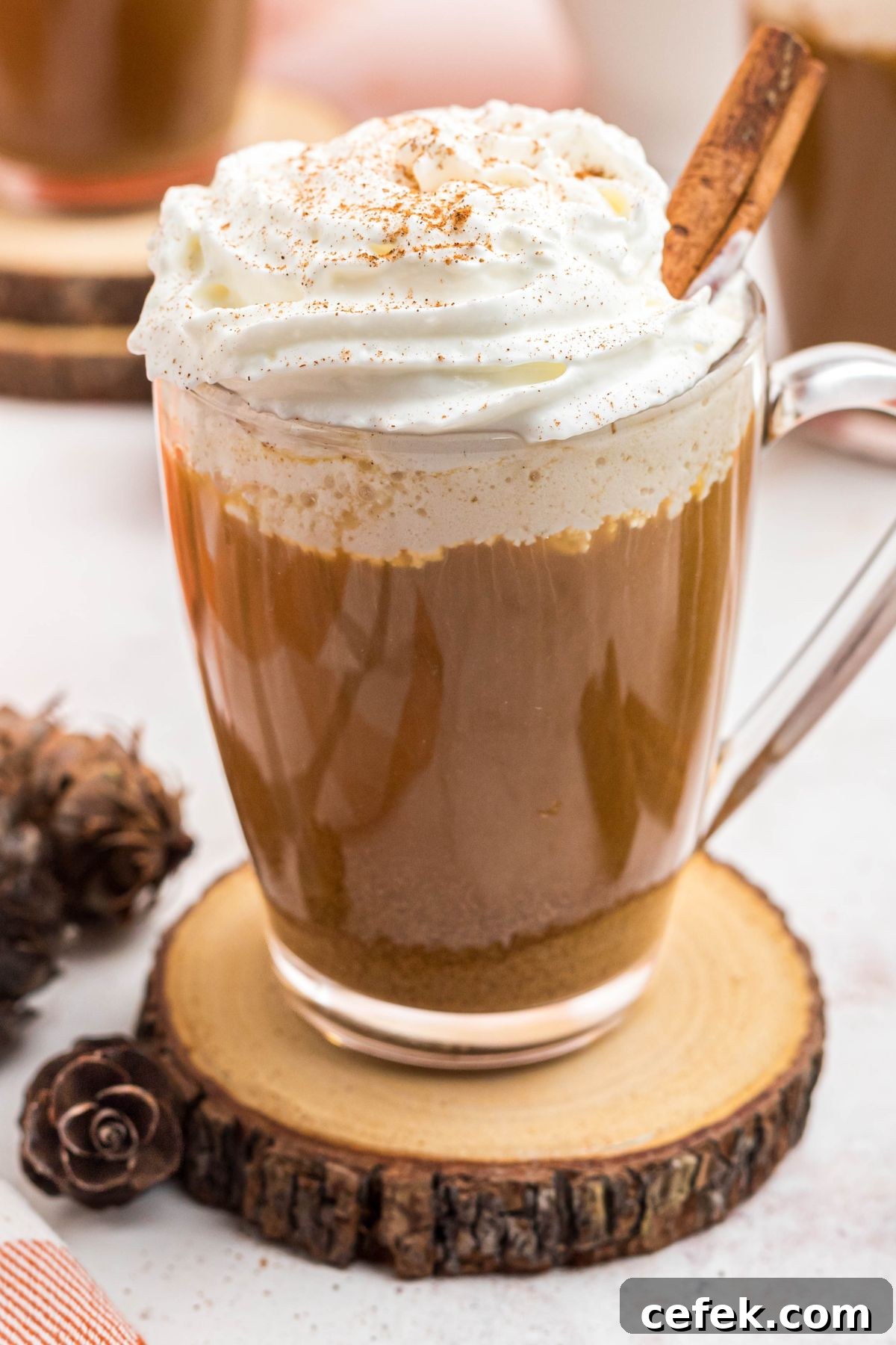 Cup of coffee topped with whipped cream and a cinnamon stick, in front of white pot, on a marble board.