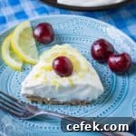 Close up shot of a slice of lemon pie garnished with a whole cherry on a blue plate with a fork, lemon slices, and more cherries on the side.