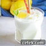 Close up side shot of frosted lemonade in a clear glass with two straws and a lemon and mint as a garnish with a blue cloth and lemons in the background.