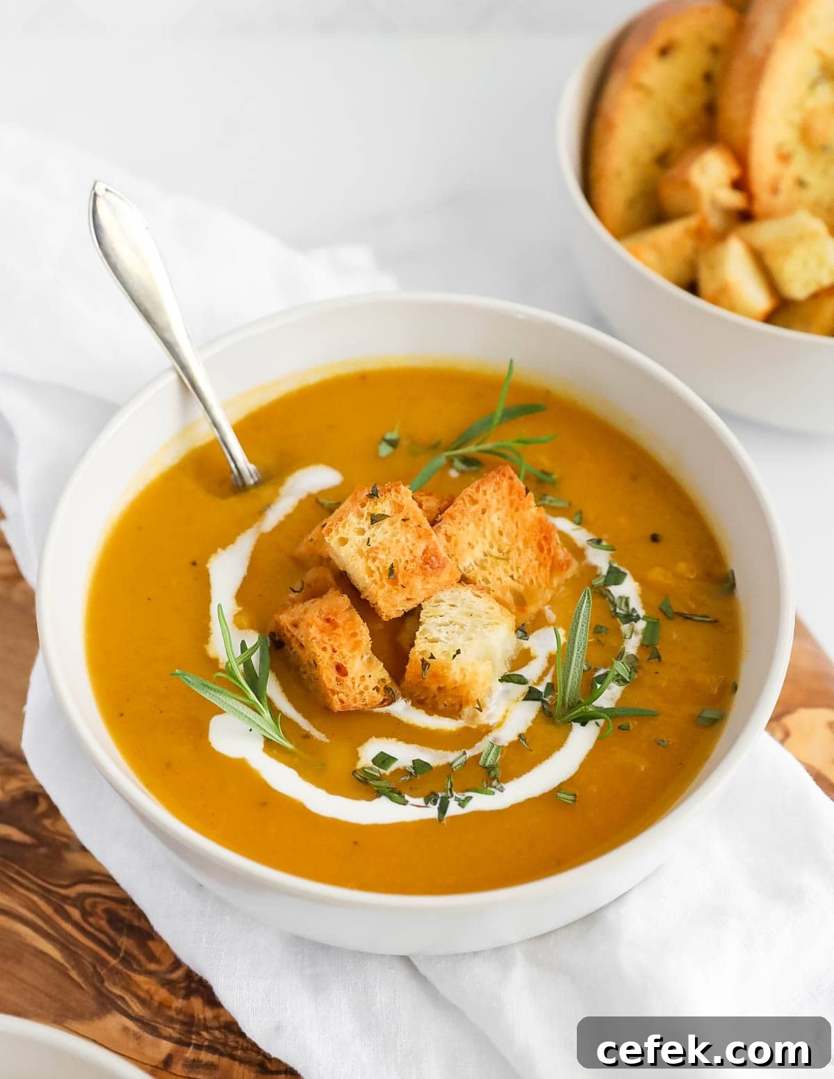 A large white bowl of orange squash soup with croutons, herbs, and coconut milk on top. There is a spoon sitting in the soup.