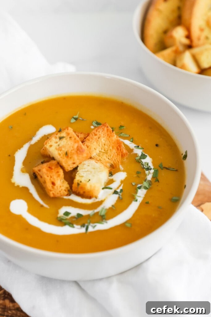 A close up image of a white bowl filled with orange soup. On top of the soup there are croutons, freshly chopped rosemary, and a drizzle of coconut milk.