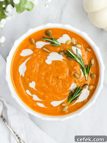 A white bowl with orange soup inside. There is white vegan crema, fresh rosemary, and pumpkin seeds garnishing the top.