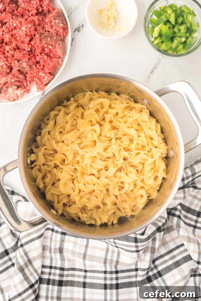 cooked egg noodles in a large pot with raw ground beef and chopped green pepper nearby