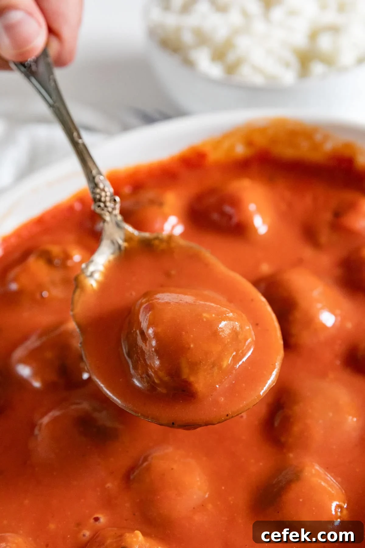A spoon holding up a porcupine meatball and sauce over the pan of them.