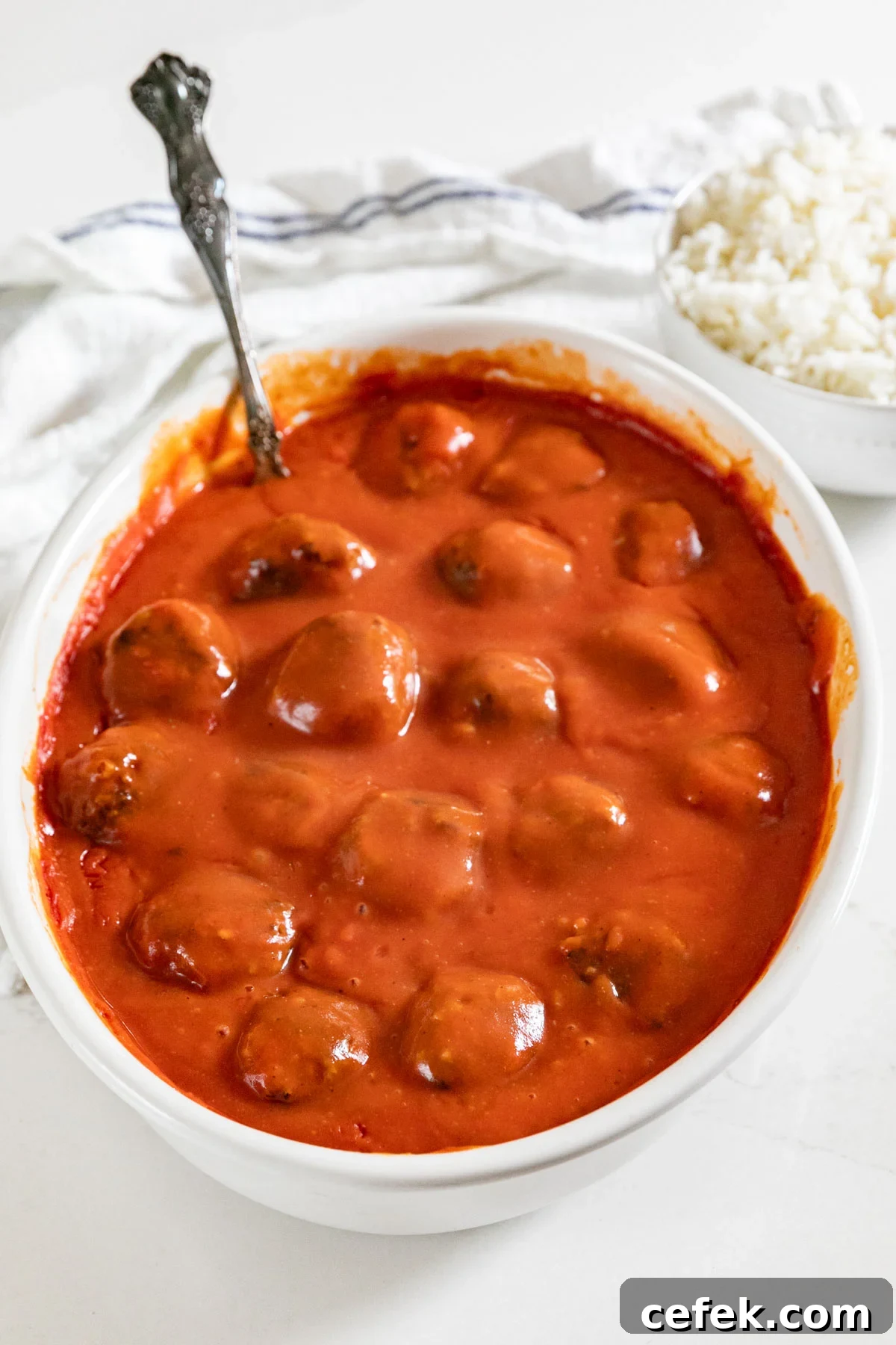 A baking pan of meatballs in tomato soup sauce with a spoon in it, a white towel and a bowl of rice in the background.