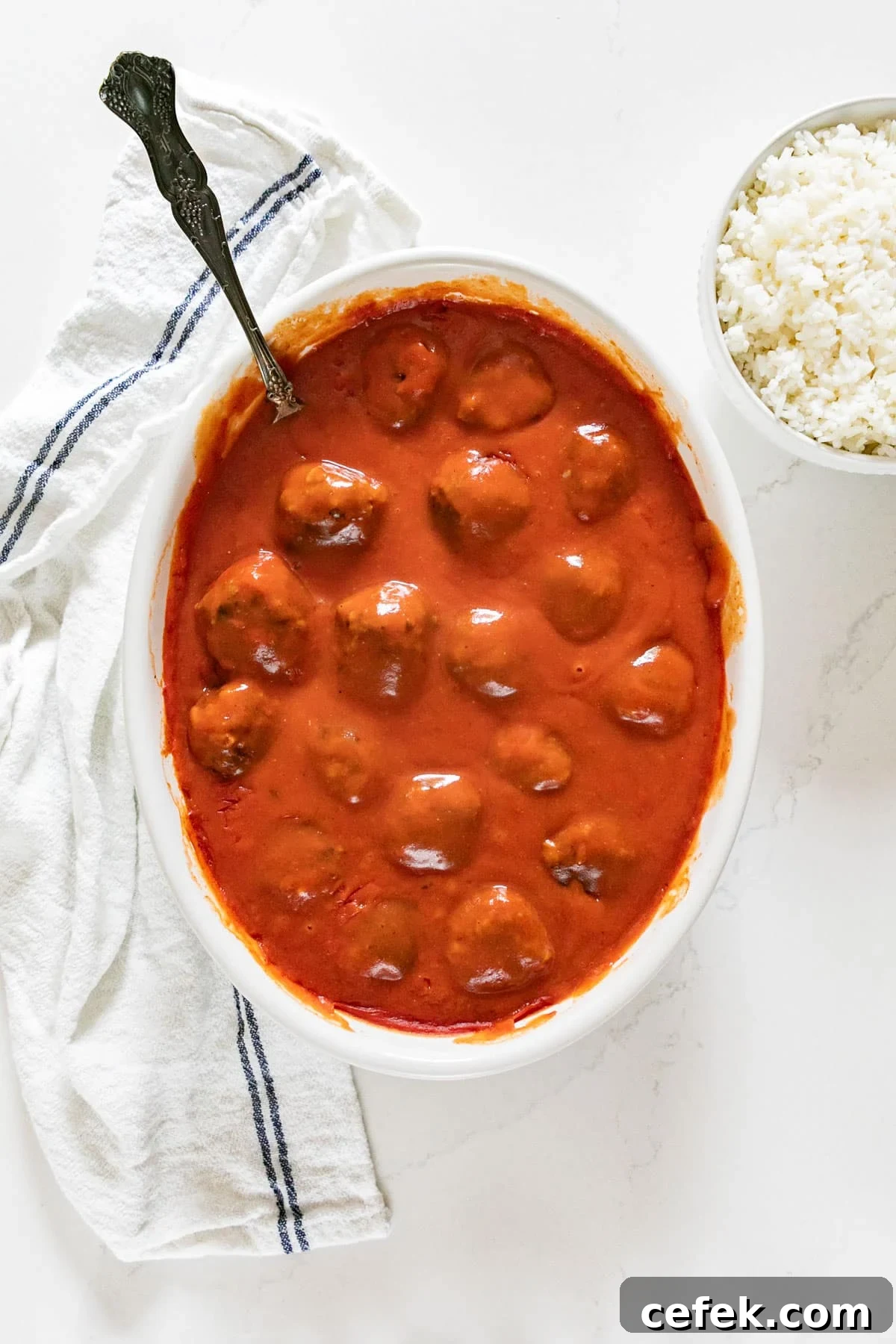 An overhead image of a pan of porcupime meatballs recipe with a spoon in it, a white and black towel and a bowl of rice on the side.