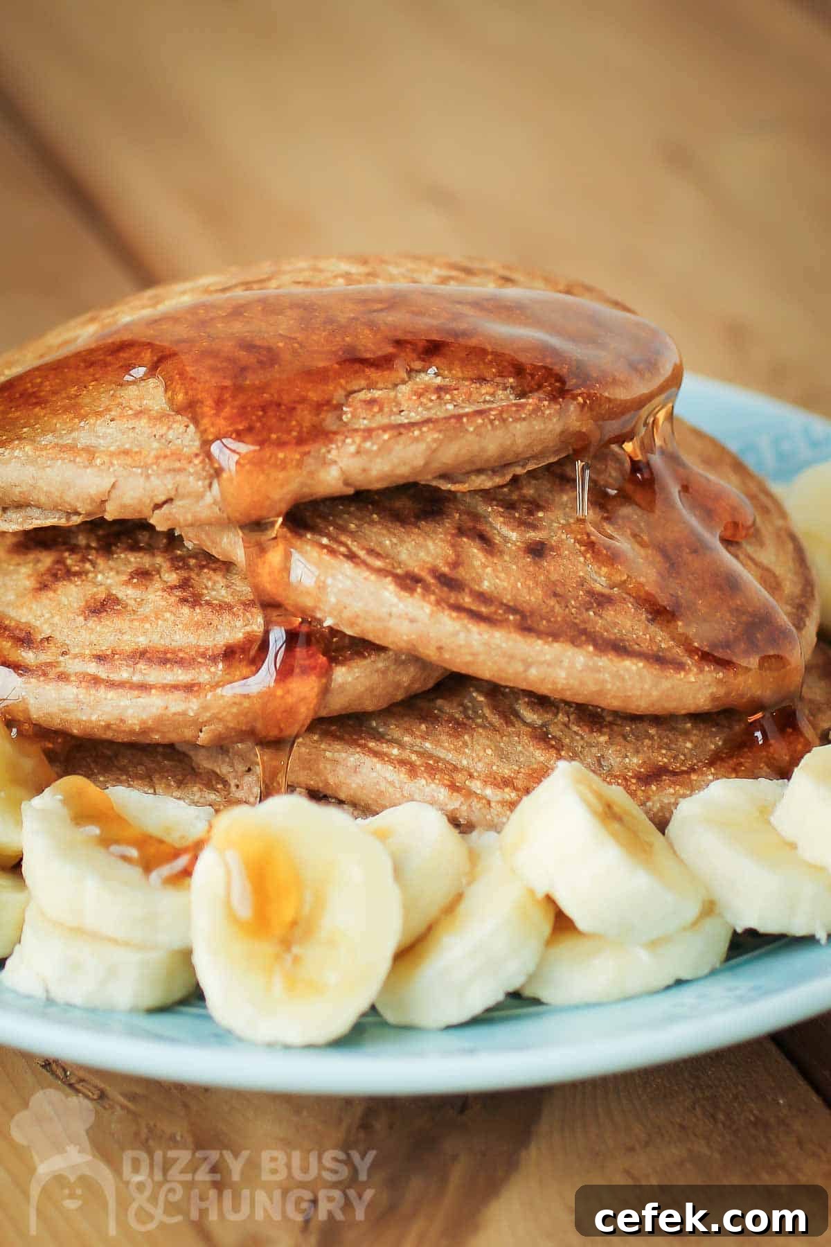 Close up side shot of multiple pancakes stacked on each other with syrup drizzled on them and bananas on the side all on a light blue plate.