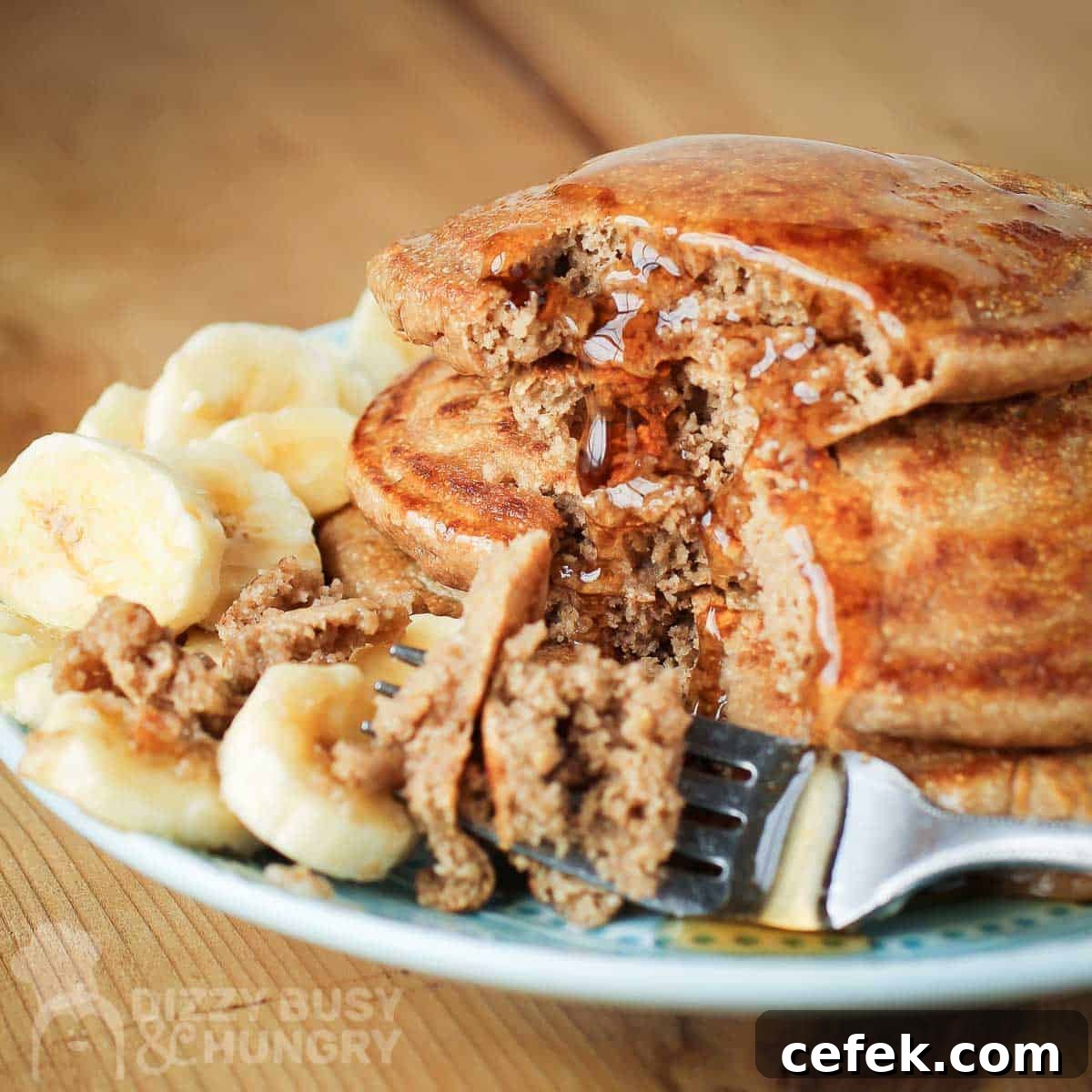 Side shot of a fork taking pieces out of multiple stacked pancakes with a syrup drizzle and sliced bananas on the side on a light blue plate.