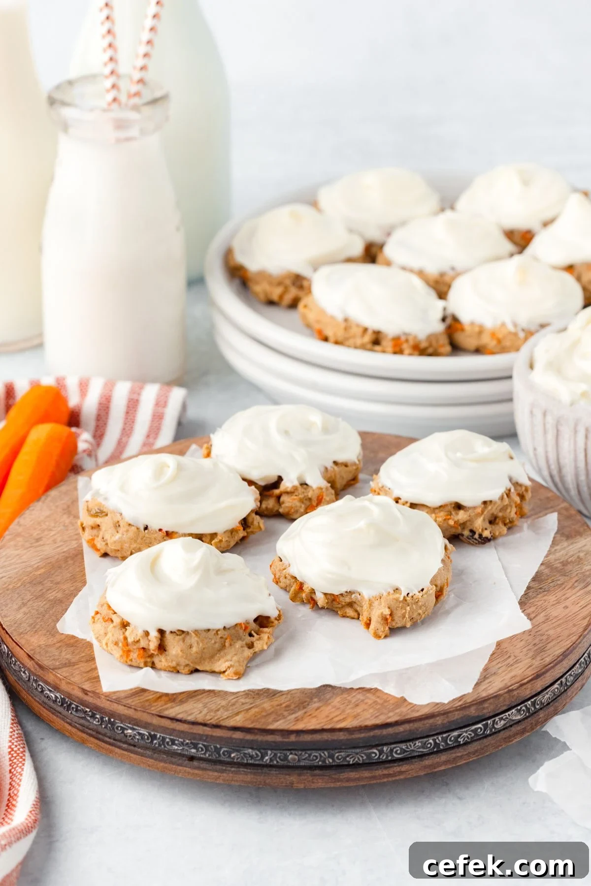 Frosted carrot cake cookies arranged on a wooden platter and a plate in the background. A bowl of cookies, two bottles of milk, a small stack of plates, and sliced carrots are also visible.