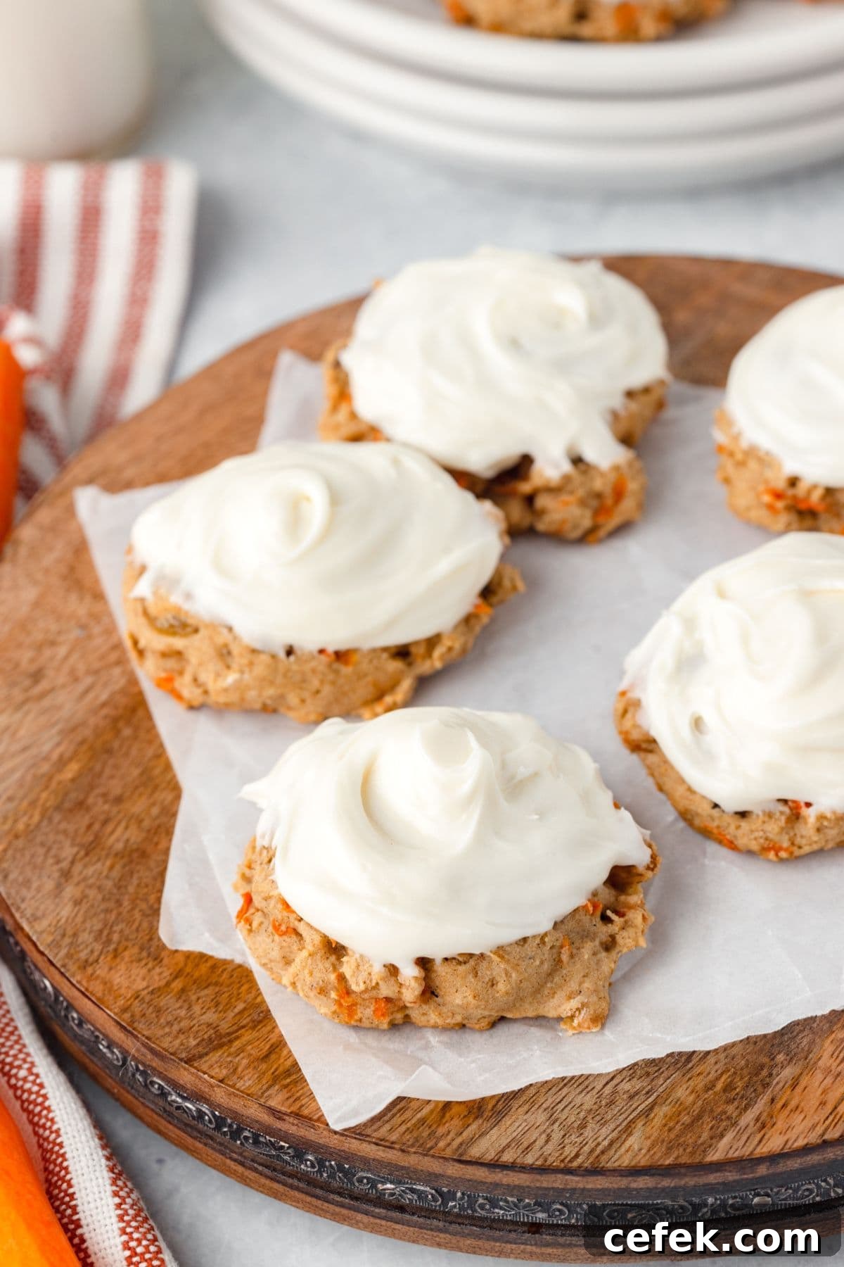 Four carrot cake cookies topped with creamy white frosting are arranged on a wooden board lined with parchment paper. A striped cloth and additional cookies can be seen in the background.
