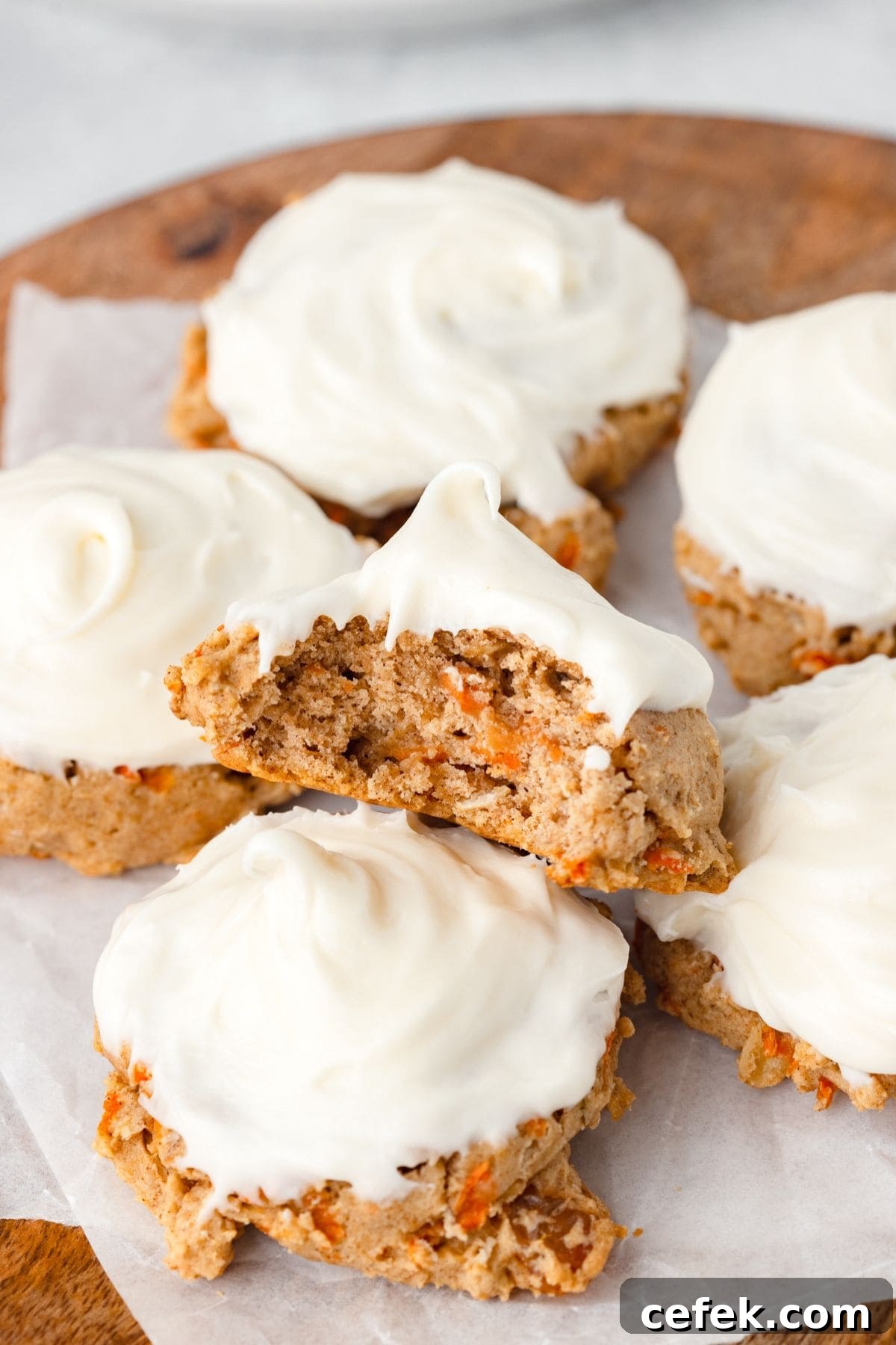 A stack of frosted carrot cake cookies on a wooden surface. One cookie is split in half, displaying the moist, spiced interior with visible pieces of carrot. The cookies are topped with smooth cream cheese frosting.
