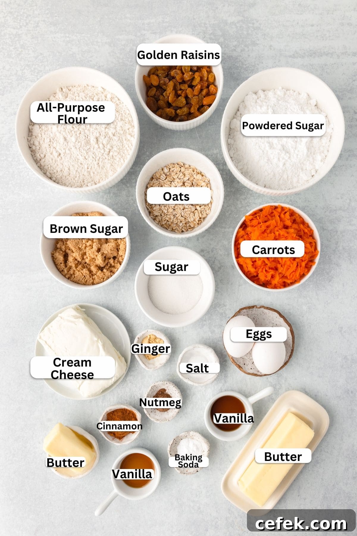 Top-down view of various baking ingredients in separate bowls: golden raisins, powdered sugar, all-purpose flour, oats, brown sugar, granulated sugar, shredded carrots, cream cheese, ginger, nutmeg, salt, vanilla, eggs, baking soda, and butter.
