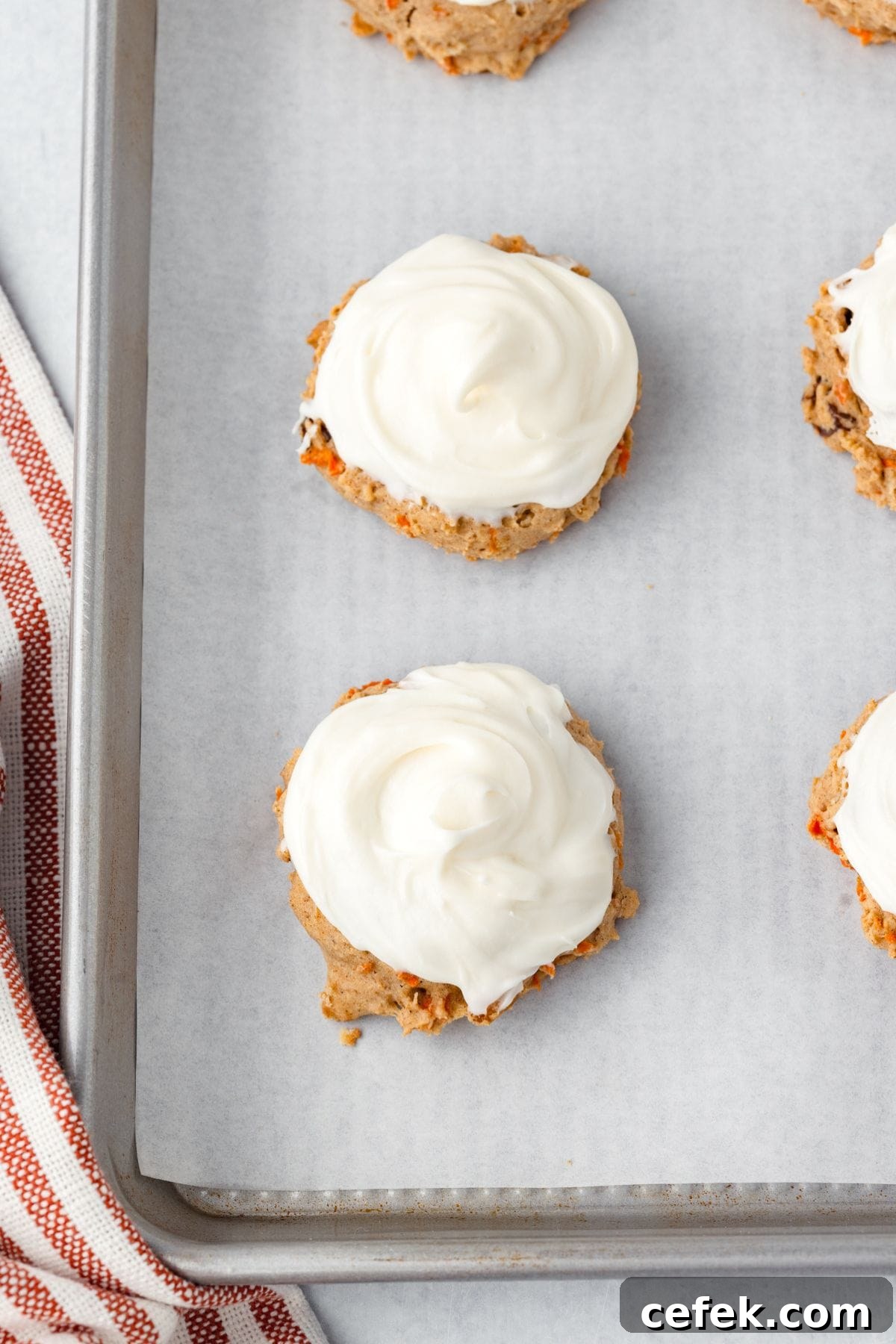 Carrot Cake Cookies with white cream cheese frosting on a parchment-lined baking sheet, next to a red-striped towel.