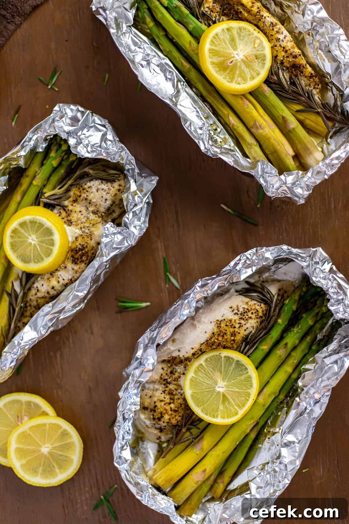 Three foil packets with baked chicken breast with asparagus spears, rosemary sprig, and lemon slice.