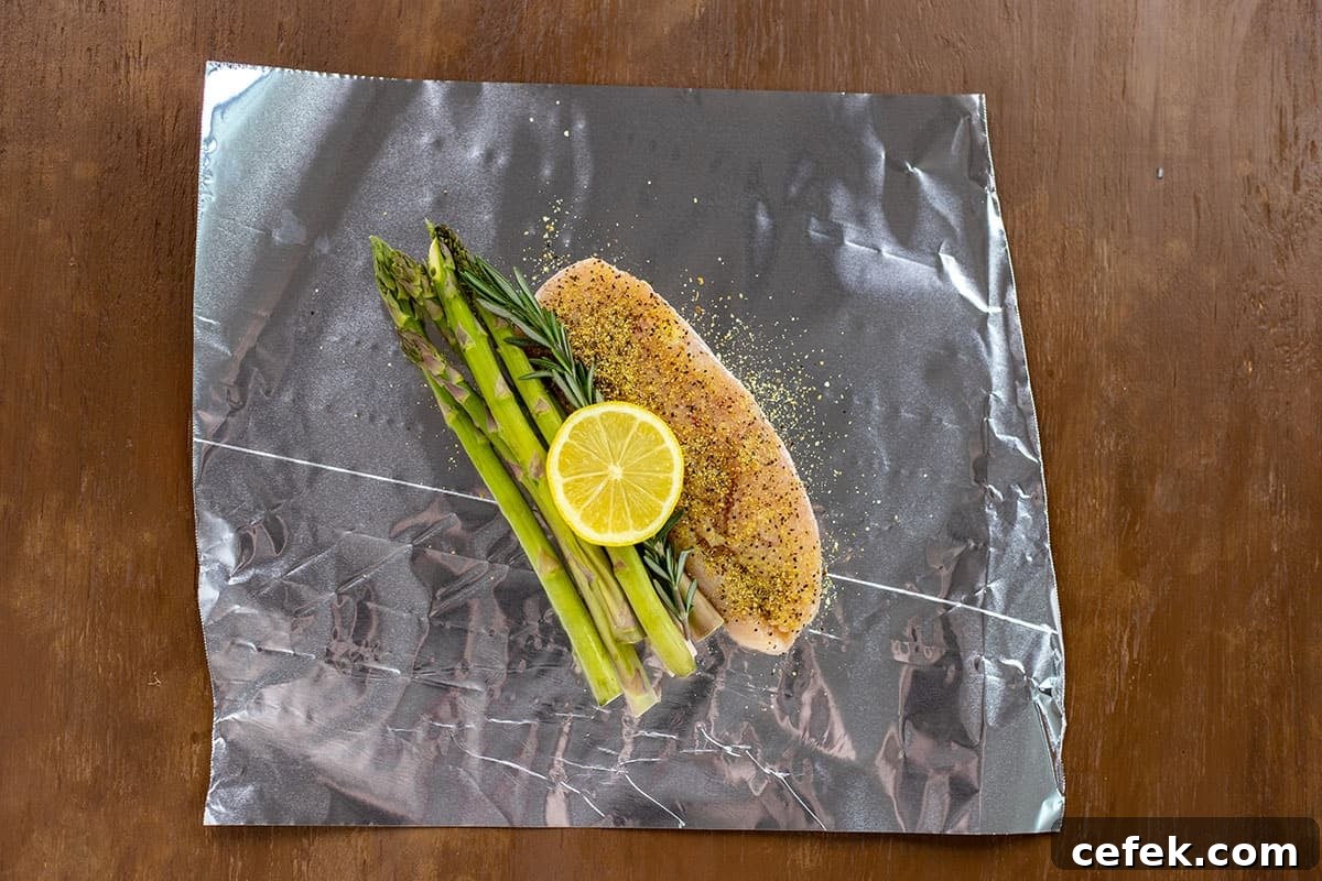 Uncooked seasoned chicken breast with asparagus, rosemary, and lemon slice on sheet of foil.