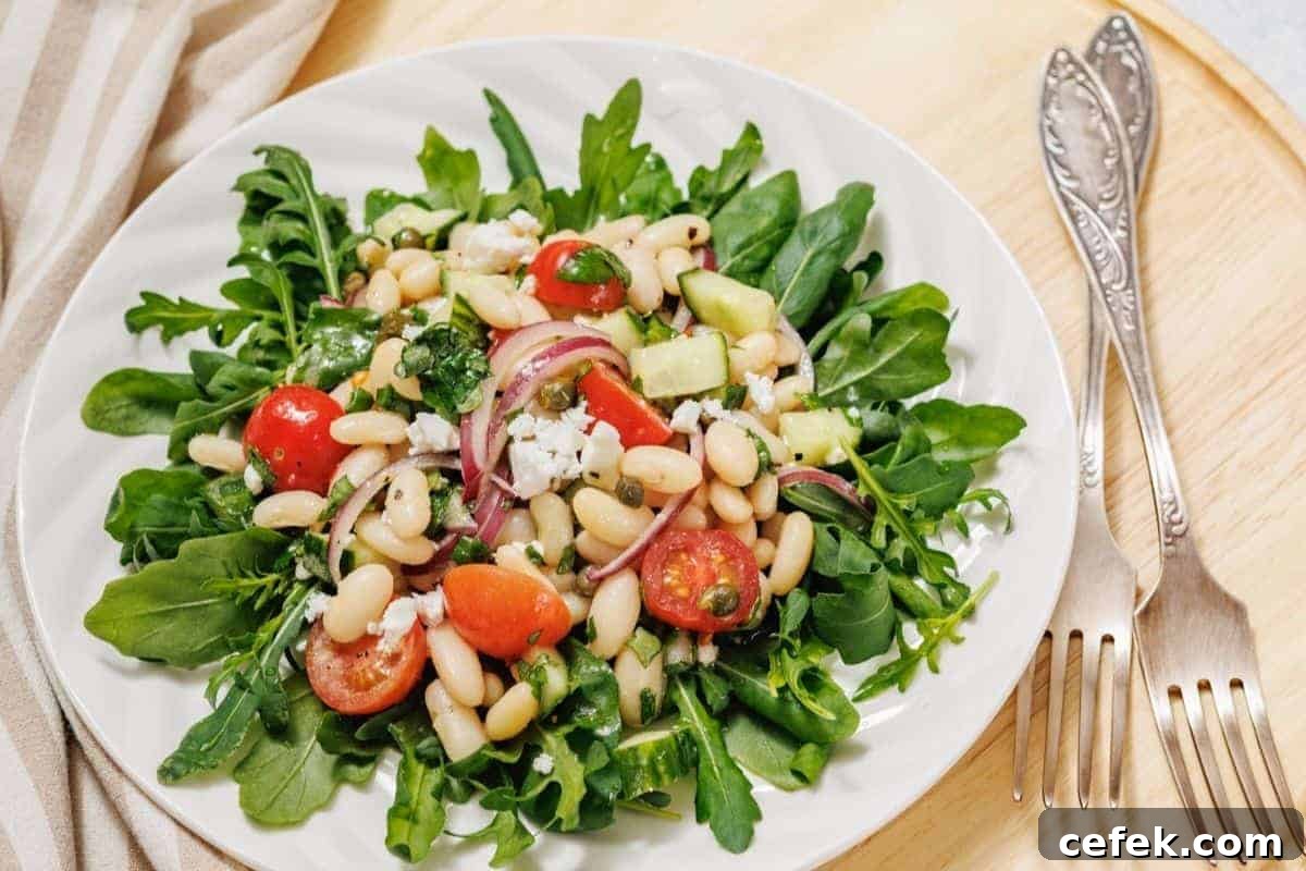A fresh White Bean Salad on a white plate featuring arugula, white beans, cherry tomatoes, sliced red onions, avocado, and cheese, placed on a wooden surface with two forks beside it.