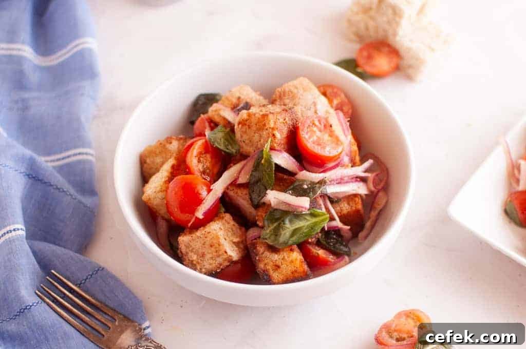 A white bowl filled with panzanella salad, featuring bread cubes, cherry tomatoes, basil leaves, and sliced red onions on a white surface.