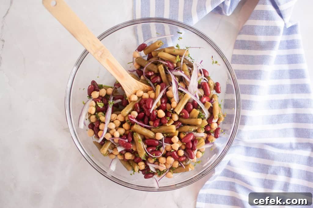 A glass bowl filled with a Three Bean Salad including kidney beans, chickpeas, green beans, and sliced red onion, with a wooden spoon and a striped cloth nearby.