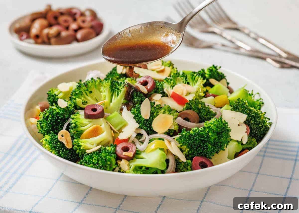 A bowl of Italian broccoli salad with sliced olives, almonds, and vegetables is being drizzled with dressing from a spoon.