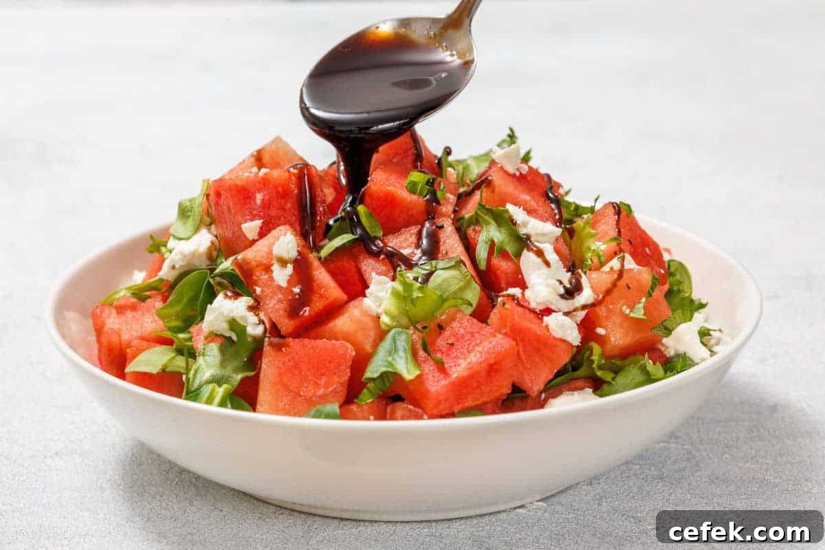 A spoon drizzles balsamic glaze over a salad with watermelon cubes, crumbled feta, and arugula in a white bowl on a light surface.