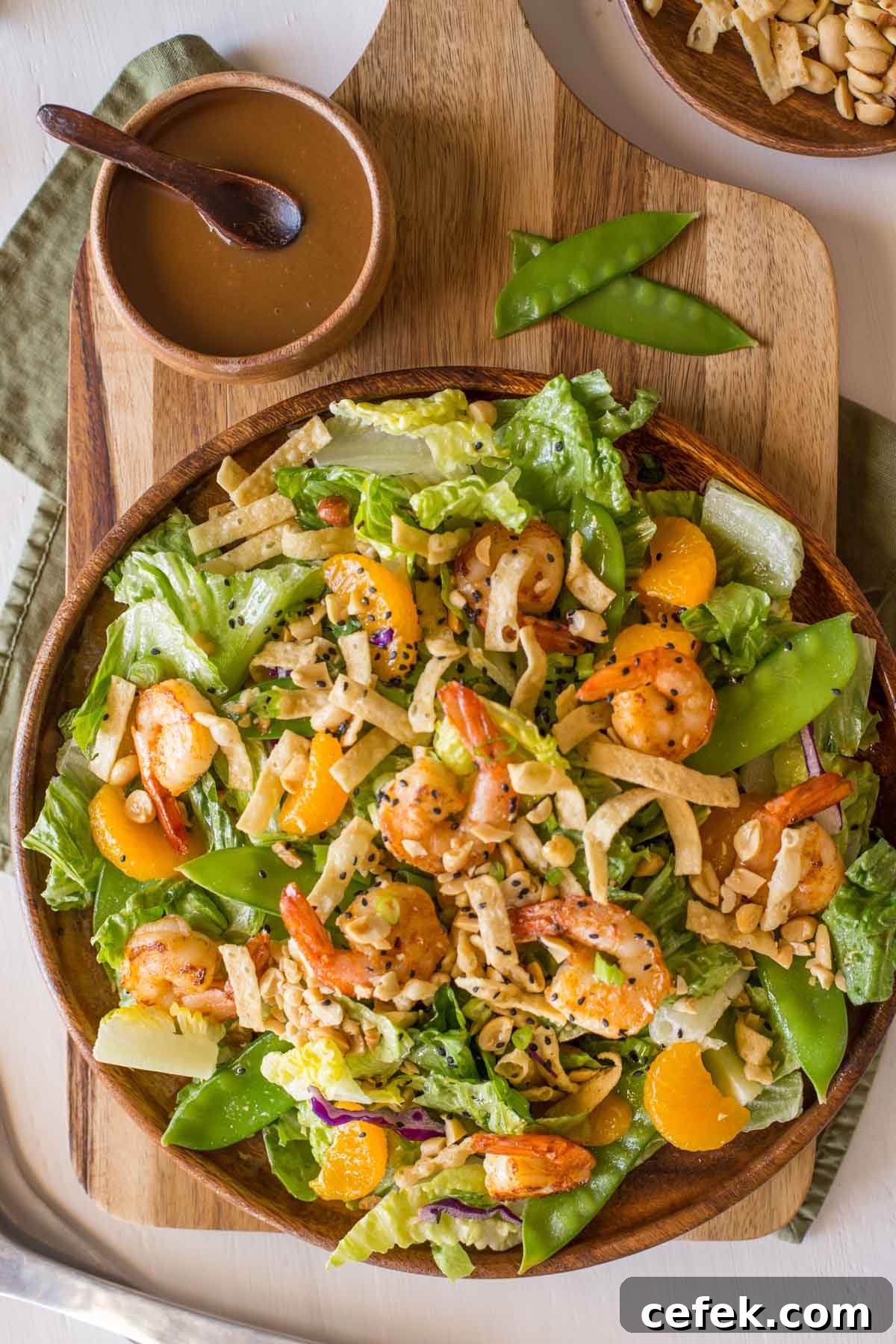 Shrimp Salad on a large serving plate, sitting next to a small wood bowl of the Spicy Peanut Dressing and a bowl of crispy wonton strips and peanuts. 