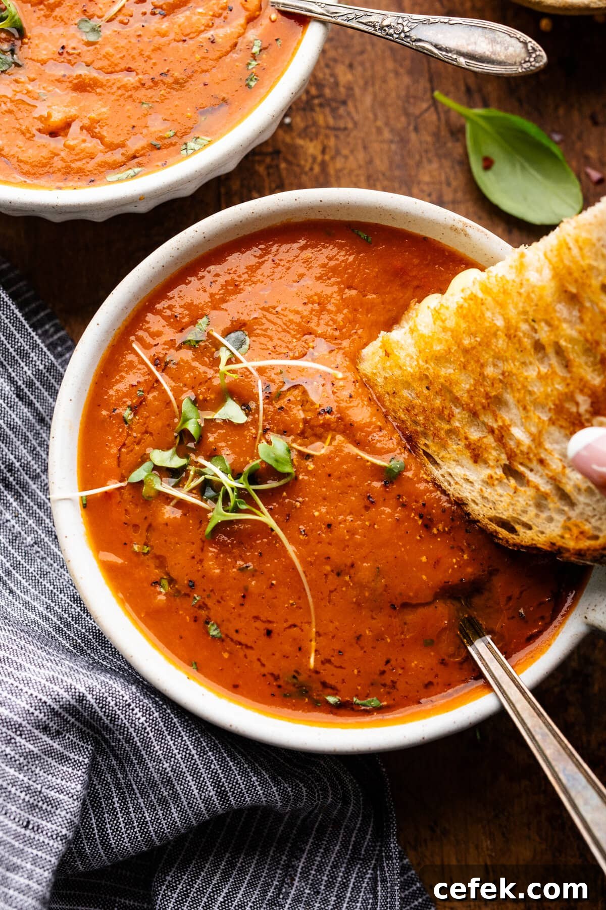 Grill cheese being dipped into a white bowl with red tomato basil soup in it. The bowl of soup is arranged on a wooden background with a dark napkin and fresh herbs displayed around it.