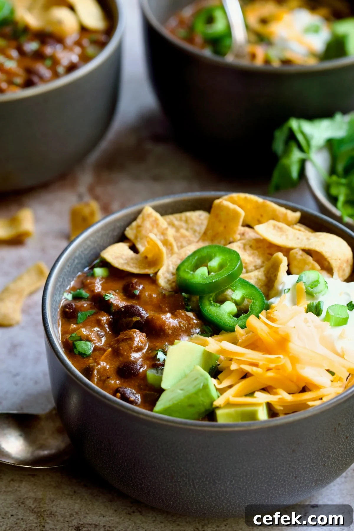 One serving of Beef and Chorizo Chili in gray bowl garnished with avocado, cheddar cheese, sour cream, jalapeno slices and corn chips.