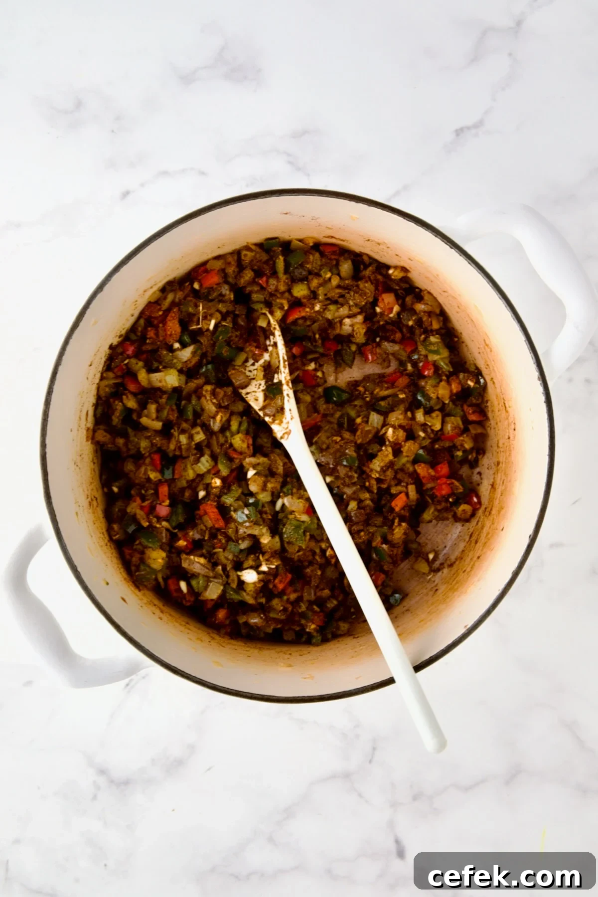 Spices stirred into cooked onions, celery, and peppers in white enamel Dutch oven with white mixing spoon.