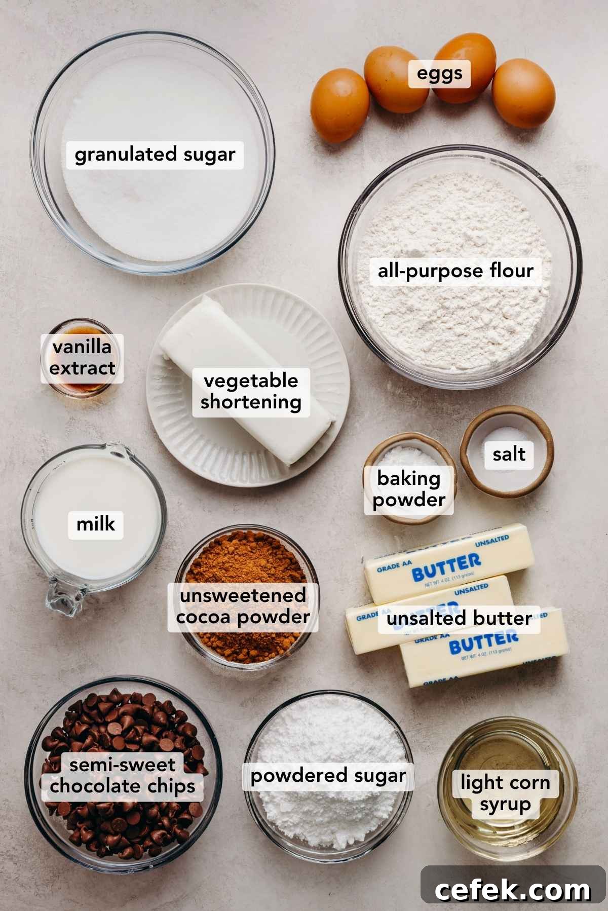 Classic Four-Egg Yellow Sheet Cake with Chocolate Frosting 5 Ingredients in various size bowls labeled as granulated sugar, eggs, all purpose flour, vanilla extract, vegetable shortening, baking powder, salt, milk, unsweetened cocoa powder, unsalted butter, semi sweet chocolate chips, powdered sugar, and light corn syrup on a white marbled background.
